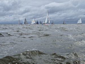 Viele Segelboote im Wasser. Das Wasser ist unruhig und wellig, der Himmel ist grau und wolkenverhangen.