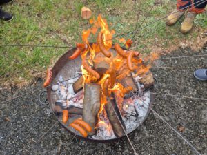 Über einer Feuerschale werden Würste am Spieß gehalten.
