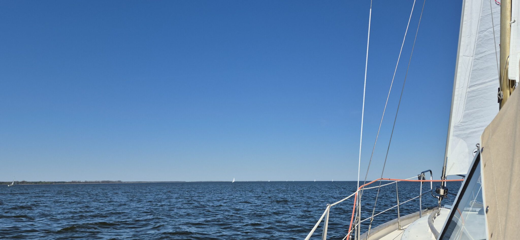 Blick von einem Segelbott auf das Wasser. Am Horizont sind einige andere Segelboote zu erkennen.